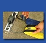 Two hands hold a blue and yellow Safcord Velcro cable cover, organizing and securing cables. A power strip with adapters is visible on the carpeted floor.